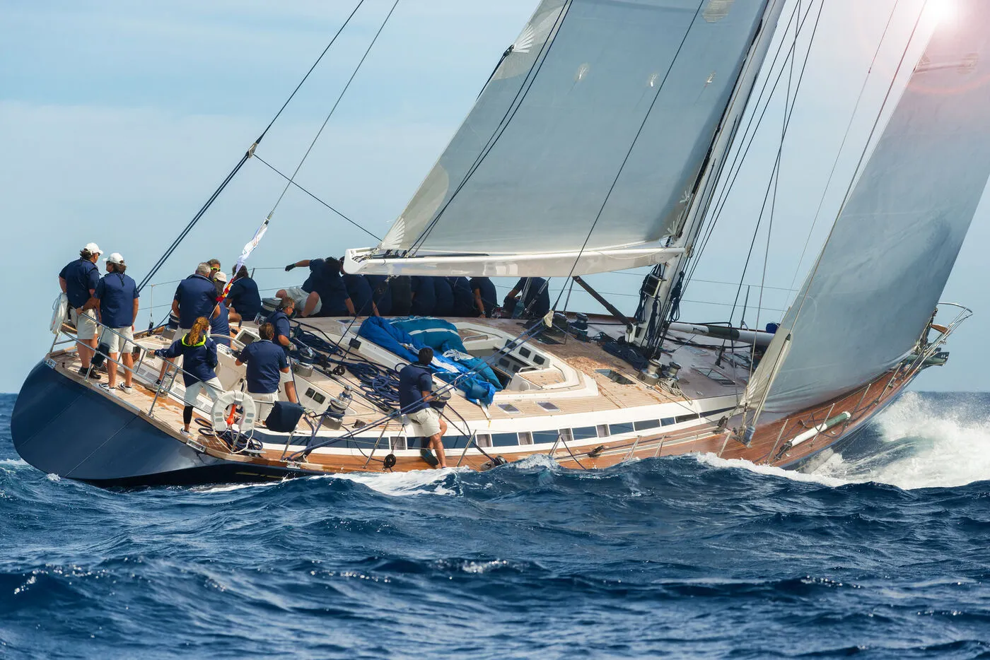 Eine Crew in dunkelblauen Shirts arbeitet an Deck einer Segelyacht, die mit vollen Segeln durch hohe Wellen fährt.